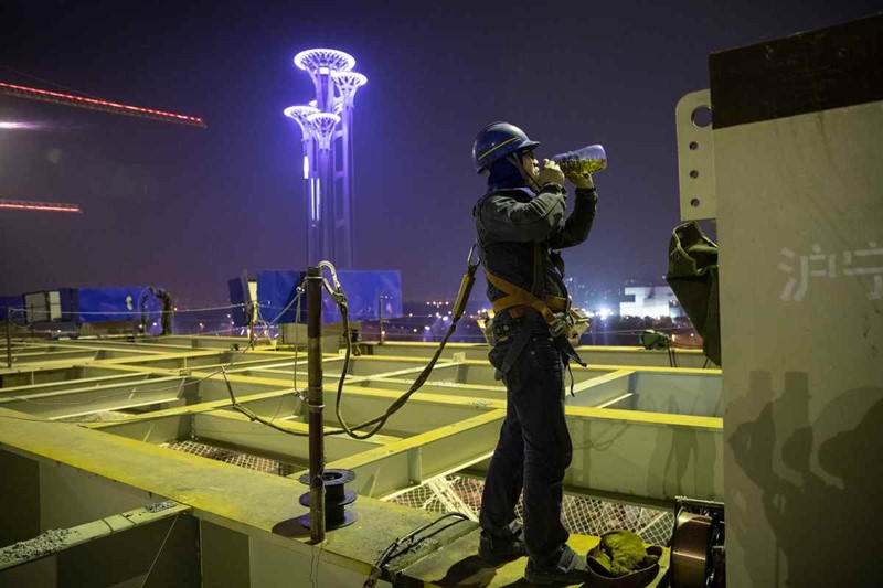 Chen Bo, un soldador que trabaja en el sitio de construcción del Centro Nacional de Convenciones, bebe agua durante su descanso el 14 de abril. La Torre Olímpica brillantemente iluminada se puede ver en el fondo. [Foto por Kuang Linhua / China Daily]