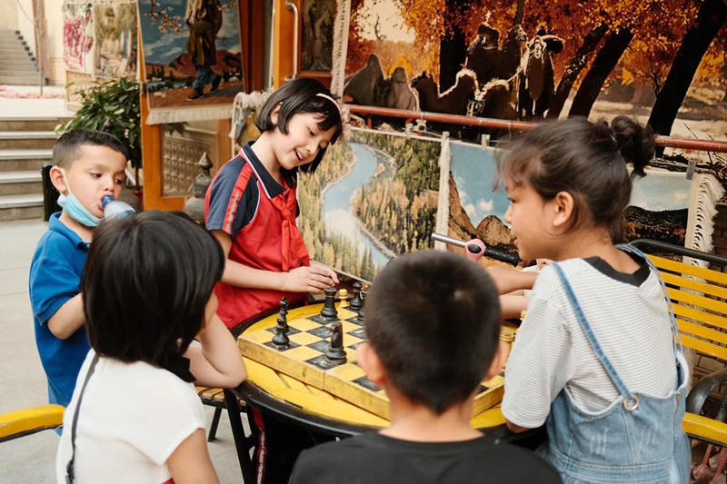 Dos ni?as Uygur juegan al ajedrez juntos después de la escuela mientras sus amigos miran el juego frente a una tienda de pintura al óleo en Tuancheng, la ruta peatonal más famosa de la ciudad de Hotan, en la región autónoma Uygur de Xinjiang, en el noroeste de China, 22 de mayo de 2021. (Pueblo en Línea / Zhang Ruohan)